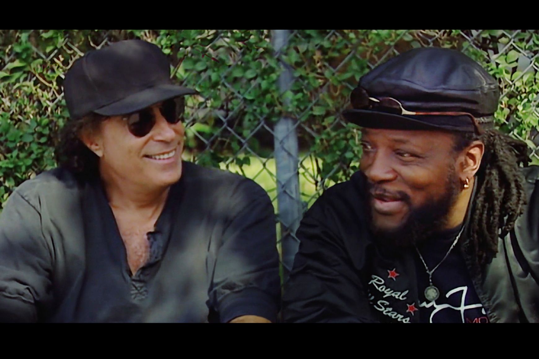 Two men sitting in front of a fence smiling 